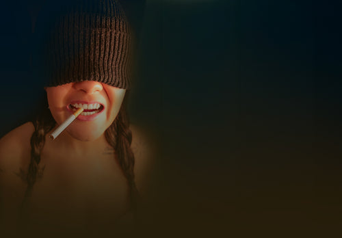 Fine art portrait of Alicea with beanie pulled over eyes, cigarette between teeth and braids, warm dramatic lighting against a dark background