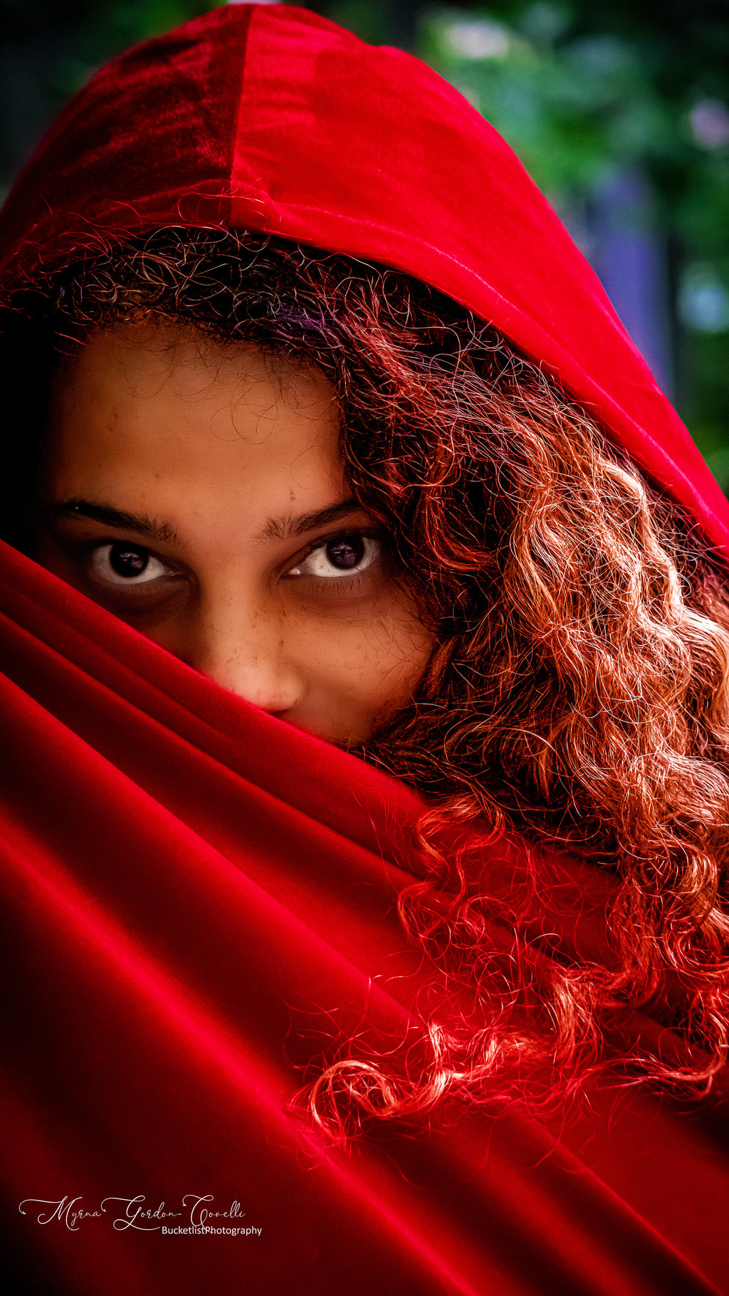 Fine art portrait of Melissa with intense expressive eyes peering over a sweeping red velvet hood, wild auburn curls framing her face against a green bokeh background