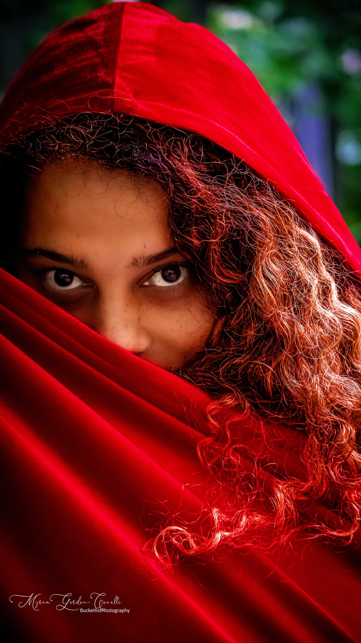 Fine art portrait of Melissa with intense expressive eyes peering over a sweeping red velvet hood, wild auburn curls framing her face against a green bokeh background