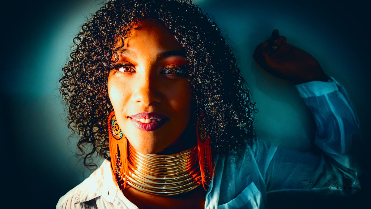 Fine art editorial portrait of Hope with natural curls, layered gold neck rings and orange beaded earrings in warm amber and teal tones