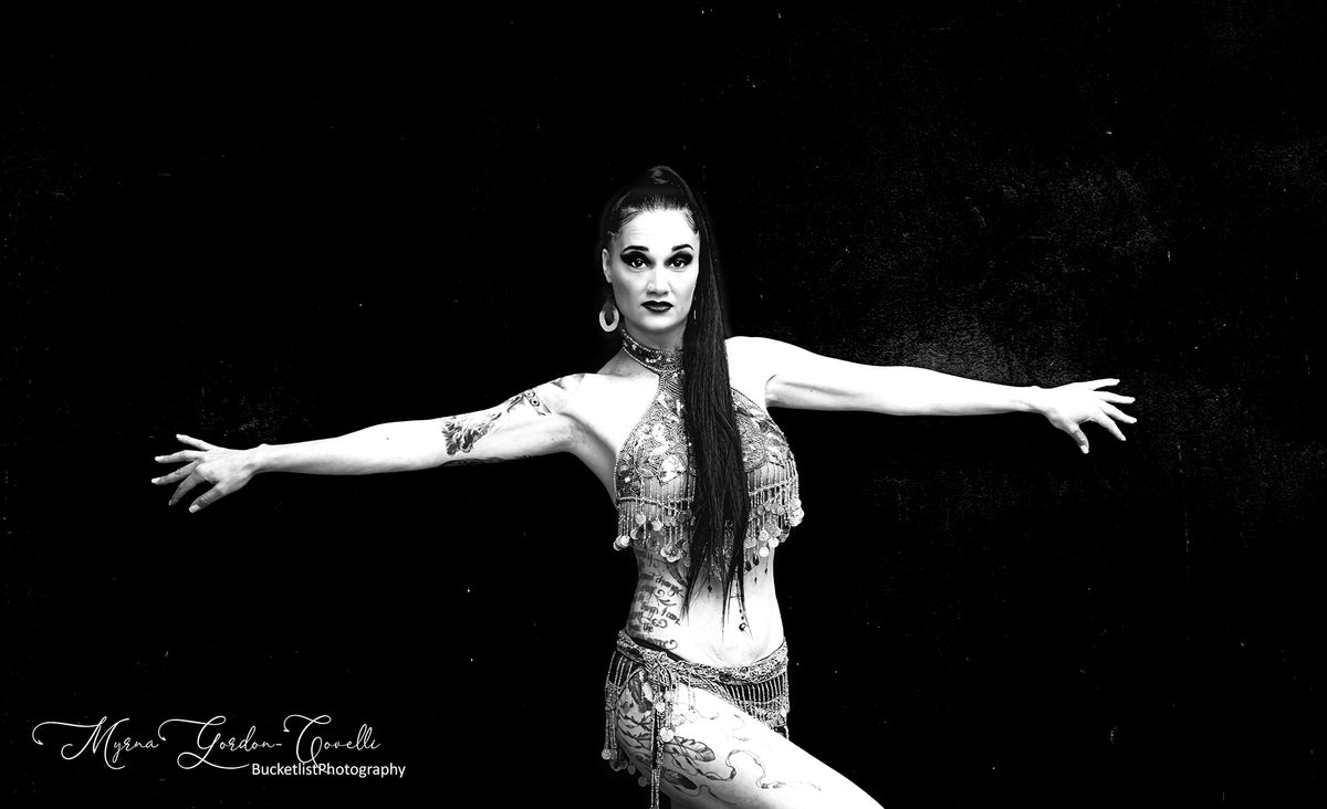 Black and white fine art portrait of Tania, a dancer with arms outstretched in ornate belly dance costume with tattoos, against a dark textured background