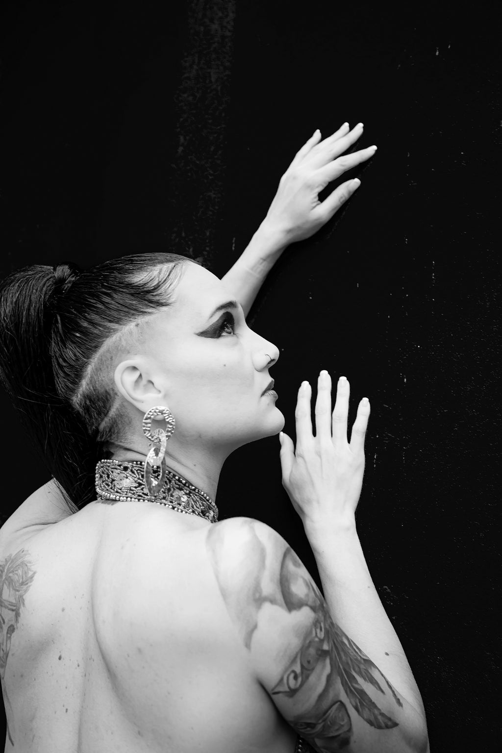 Black and white fine art portrait of Tania in sharp profile with shaved undercut, chain choker, tattoos and one hand raised against a dark textured background