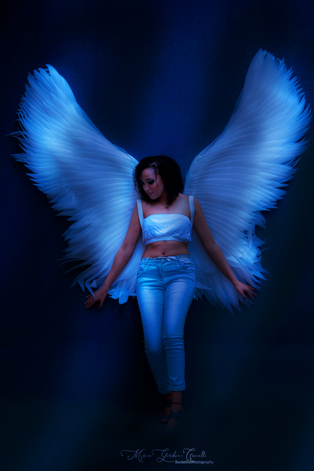 Fine art portrait of Megan with large luminous white angel wings spread wide, bathed in deep blue atmospheric light, head bowed in quiet contemplation