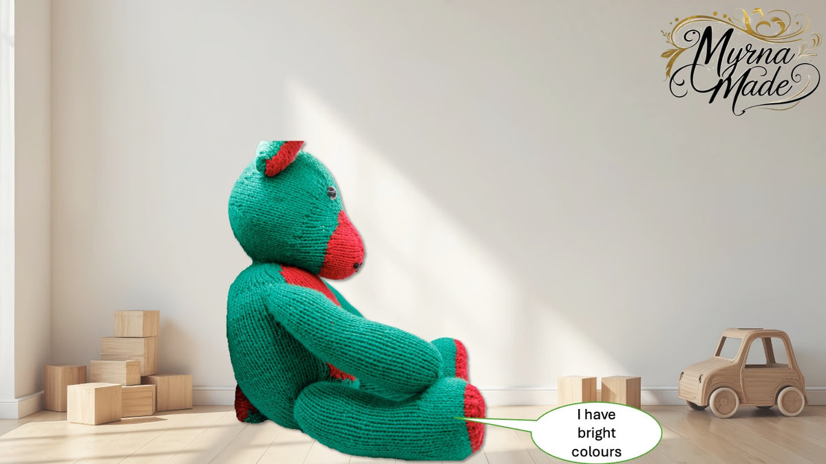 Green teddy bear with red accents sitting on a wooden floor with toys around, featuring 'Myrna Made' branding.