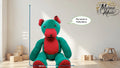 Knitted teddy bear with a speech bubble saying 'My name is Holly Berry' in a room with toys and a ruler for scale.